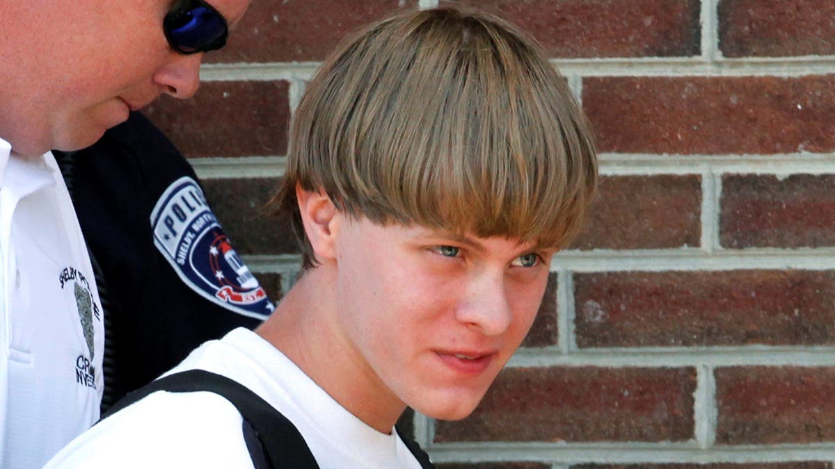 Convicted mass murderer Dylann Roof being led into a courthouse by police. He has a bowl haircut and is wearing a white T-shirt and bulletproof vest