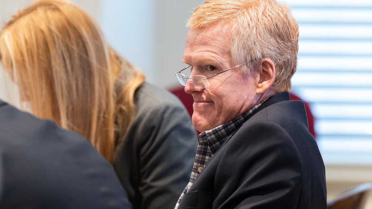 A man wearing glasses and a suit looks on inside a courtroom.