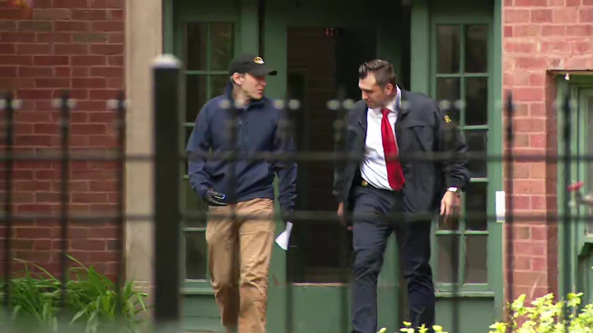 Two investigators talking outside the apartment complex, seen through a metal fence