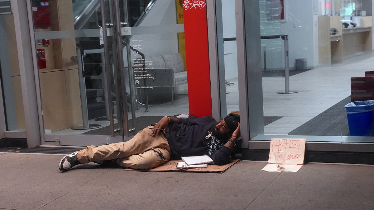 homeless man lying on sidewalk by glass front of building
