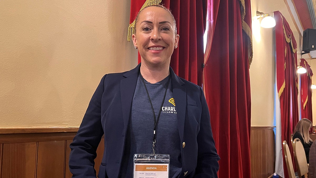 Bekah Charleston wearing a sign around her neck and a blue blazer with a matching shirt smiling.