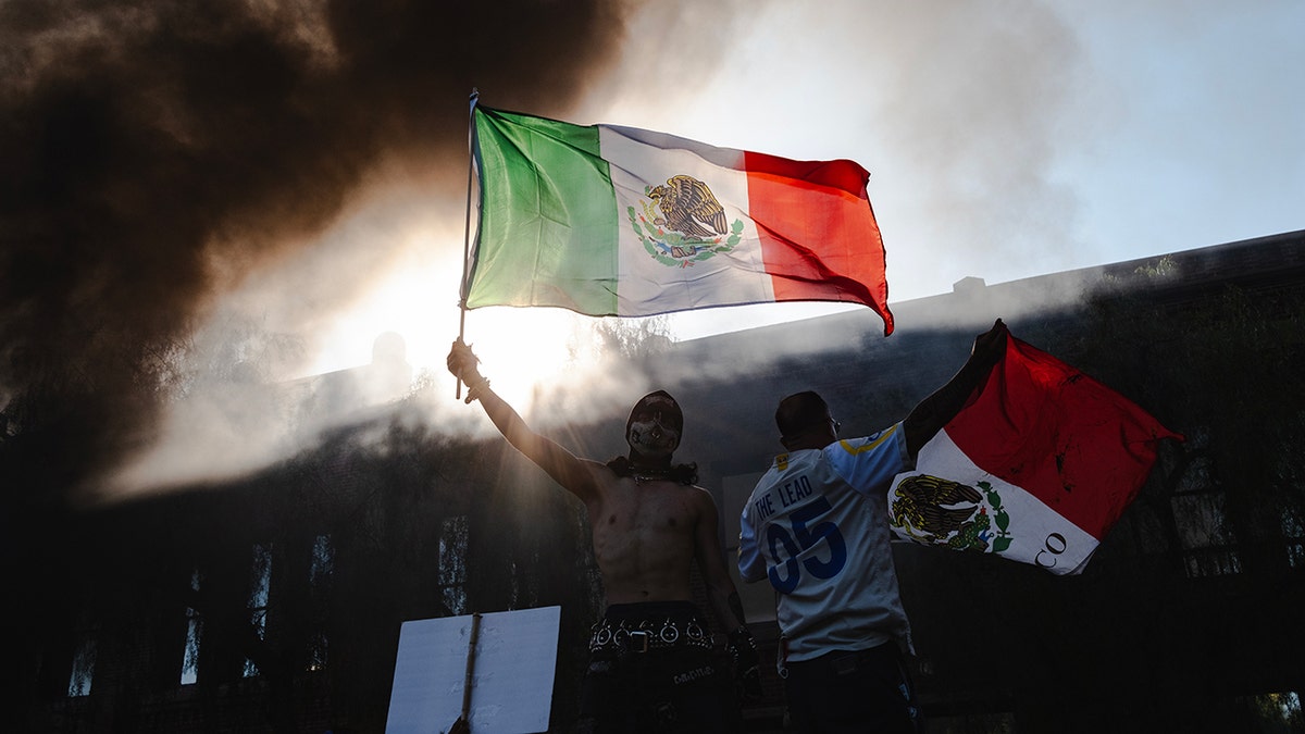 Rioter waving Mexican flag amid smoke from burning cars