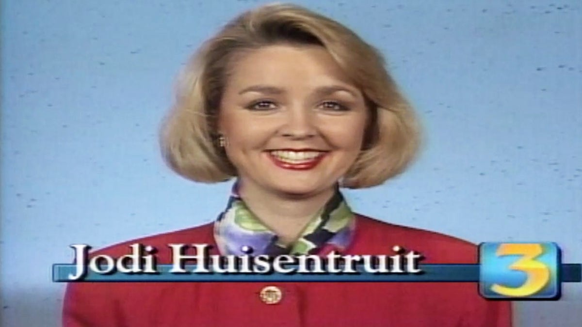 Jodi Huisentruit on the TV screen wearing a red dress and a floral scarf.