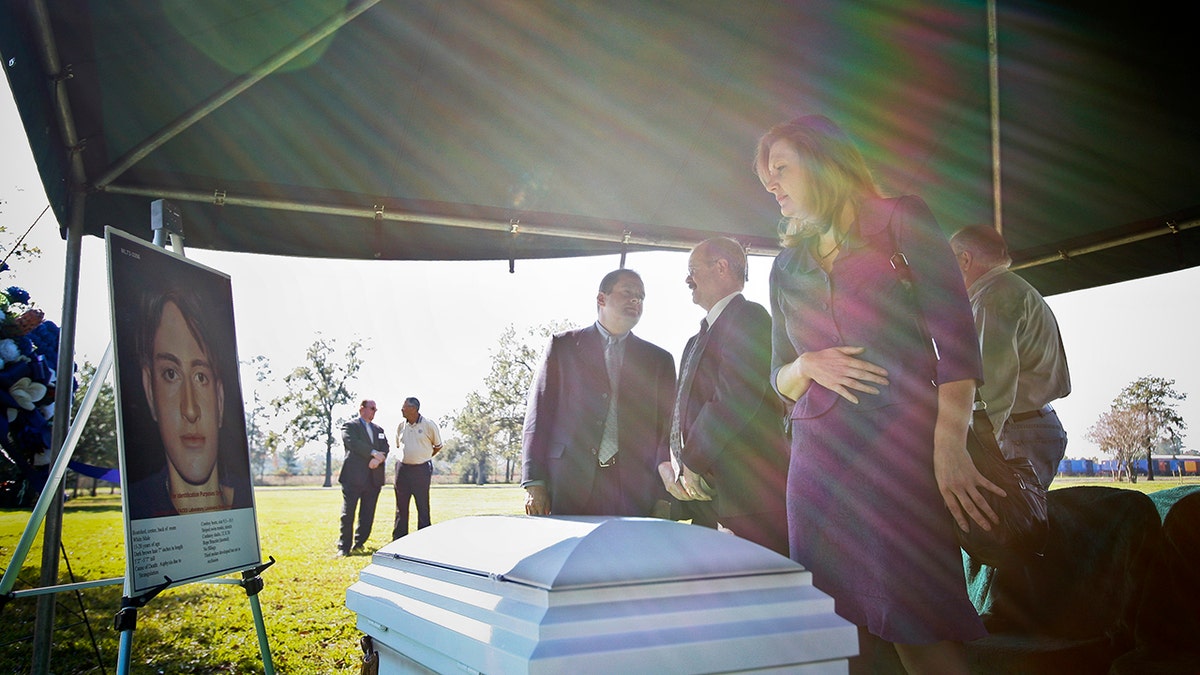 A woman looking ahead at a white casket and a photo of a murdered victim.