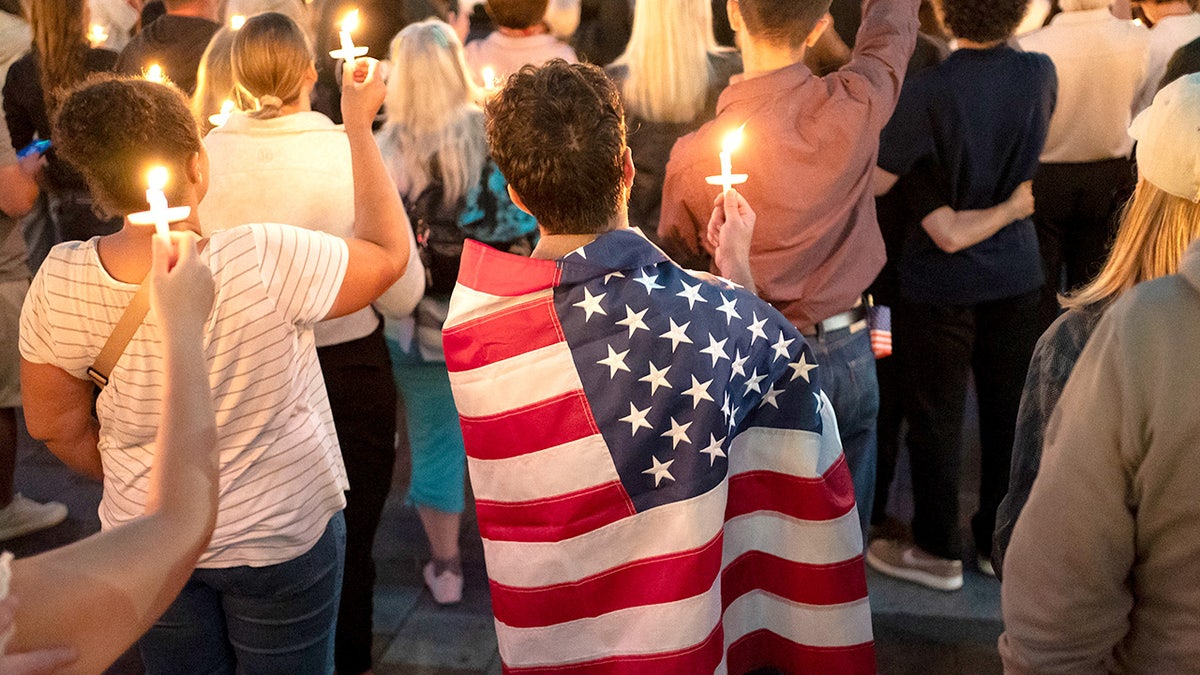 Person wearing a U.S. flag stands in a crowd of people holding candles during a vigil for Charlie Kirk.