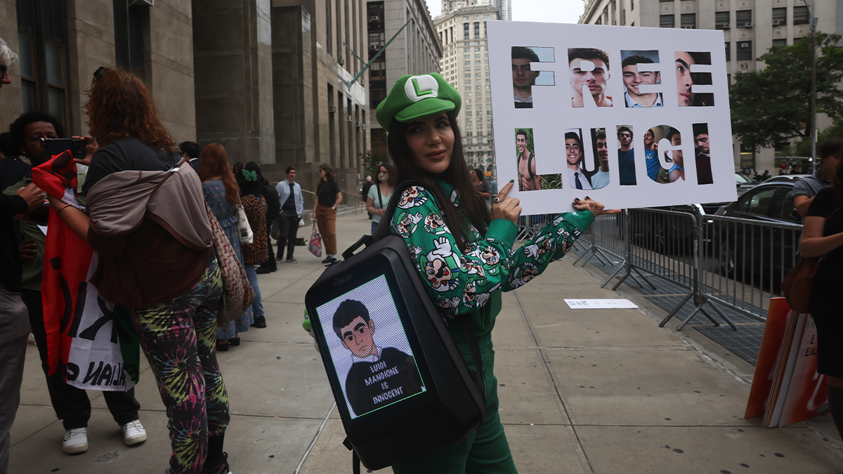 A woman dressed up as the character Luigi from "Super Mario Bros" holds a sign that says "Free Luigi" outside a hearing for suspected assassin Luigi Mangione