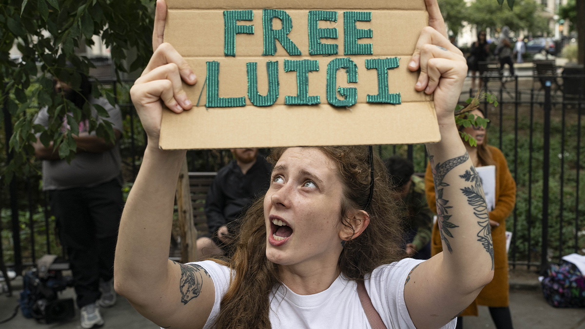 A woman with red hair wearing a white t-shirt holds up a cardboard sign that says "free luigi" in green letters