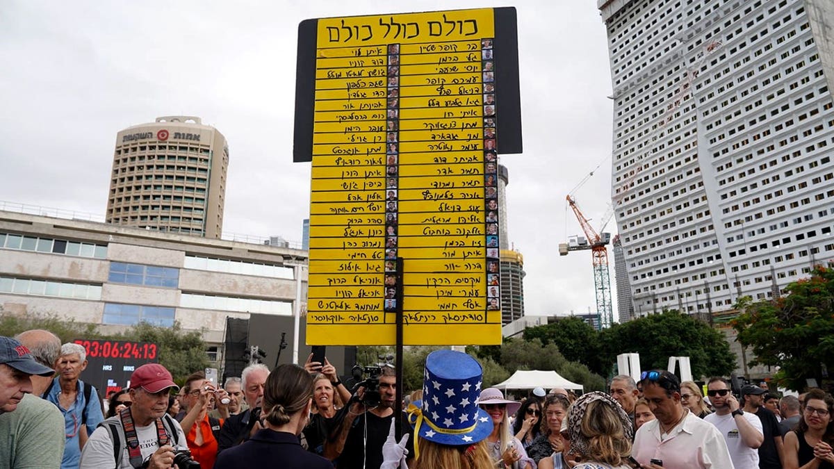 People gather in Hostages Square as a woman holds a large yellow sign listing hostages’ names in Hebrew.