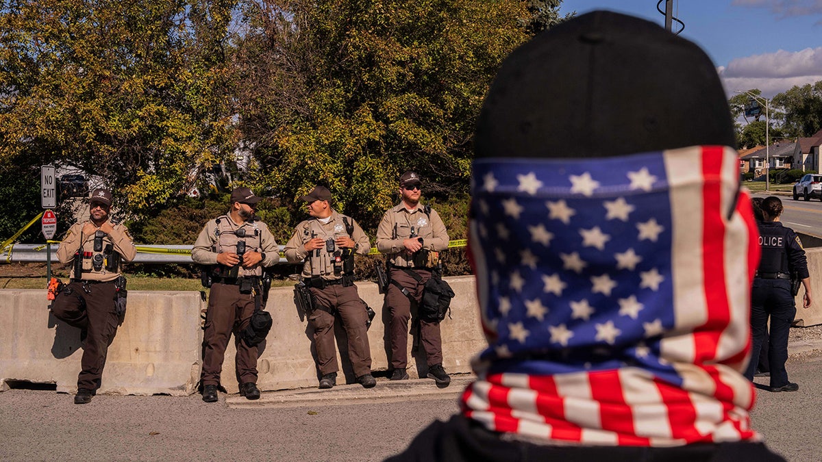 A protestor wearing an American flag face covering stands opposite Cook County Sheriffs outside of an Immigration and Customs Enforcement facility in Broadview, Ill., Saturday, Oct. 11, 2025.