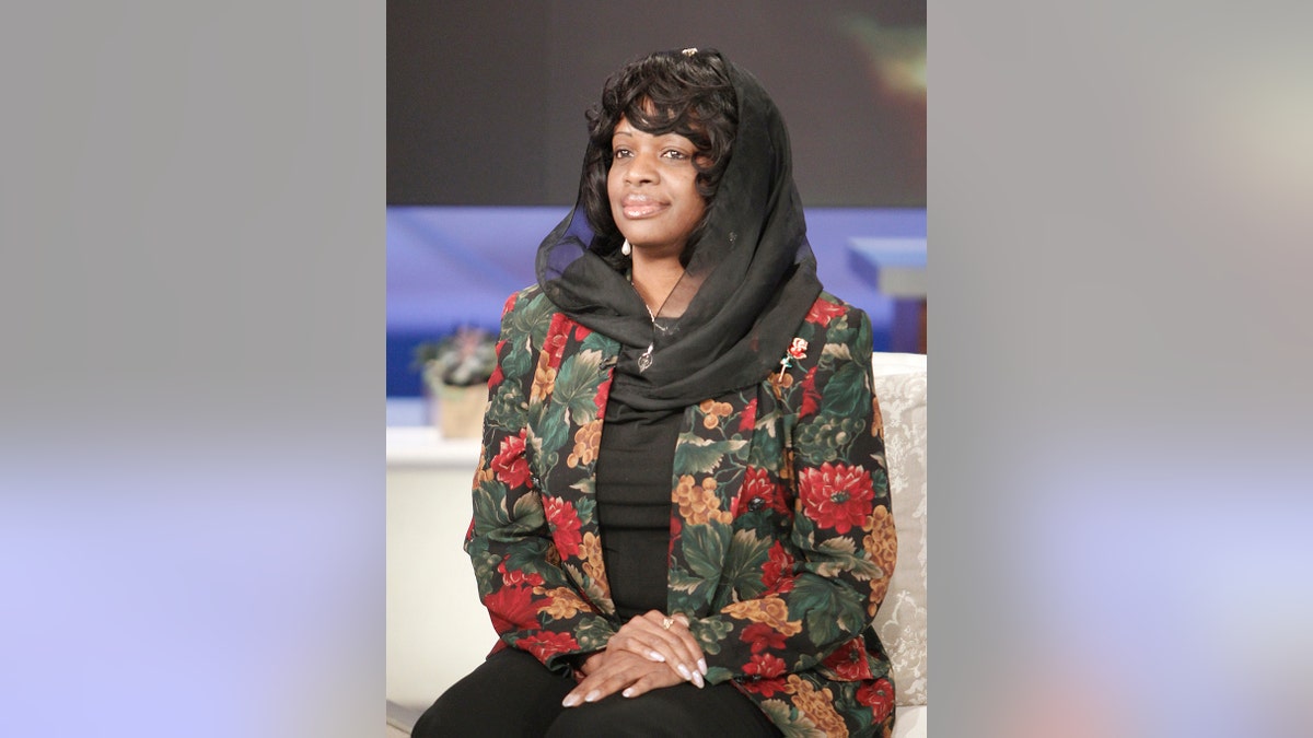 Mildred Muhammad in a floral suit looking poise and sitting on a white couch.