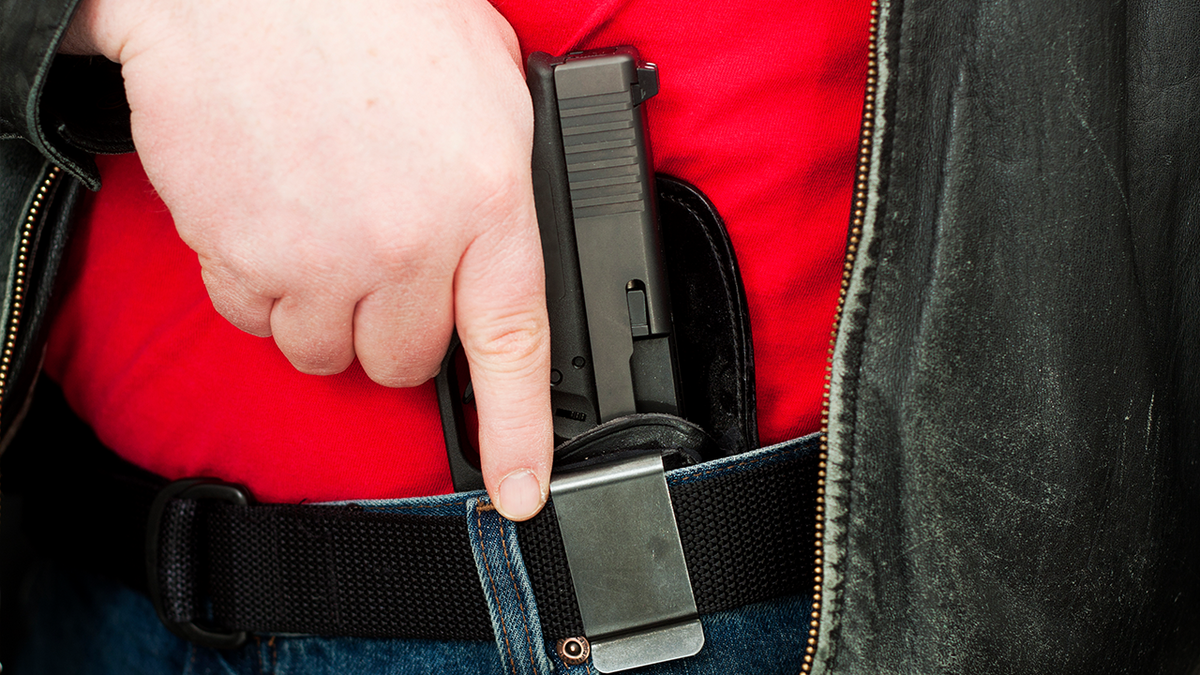 Stock image shows a man in a red t-shirt and jeans drawing a black handgun from an IWB holster