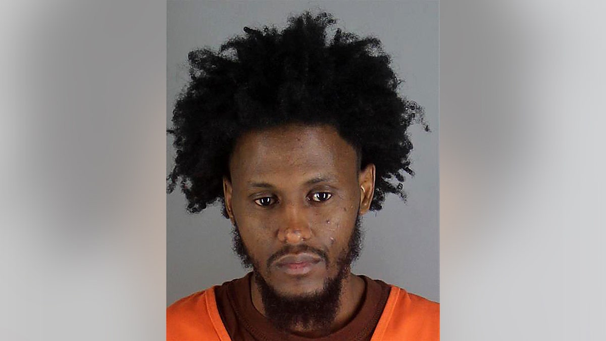 Man looks downward in a close-up with short beard and orange clothing against a plain background.